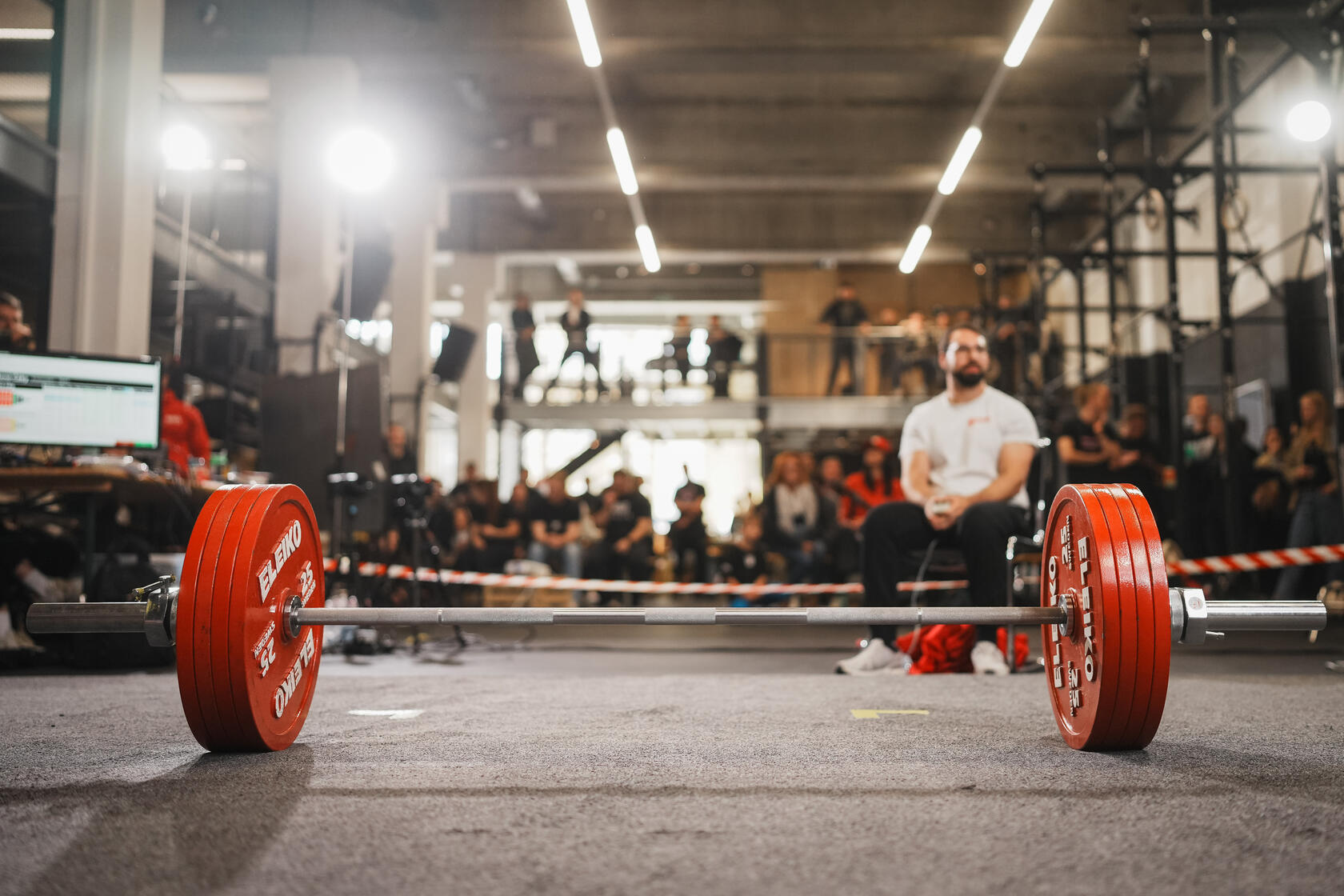 © Dedicated Sports Auf dem Bild ist eine beladenen Langhantel zu sehen. Es sind pro Seite vier rote Eleiko Gewichtsscheiben an je 25kg mit einem eisernen Verschluss festgemacht. Die Langhantel steht auf dem Teppichboden an einem Wettkampf. Im Hintergrund sind Menschen.